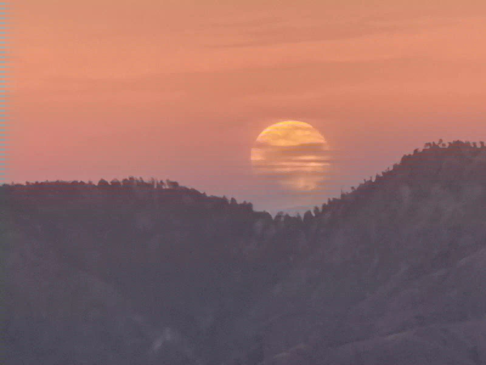 The golden Harvest Moon rising through haze into a pink sky, between two forested ridges