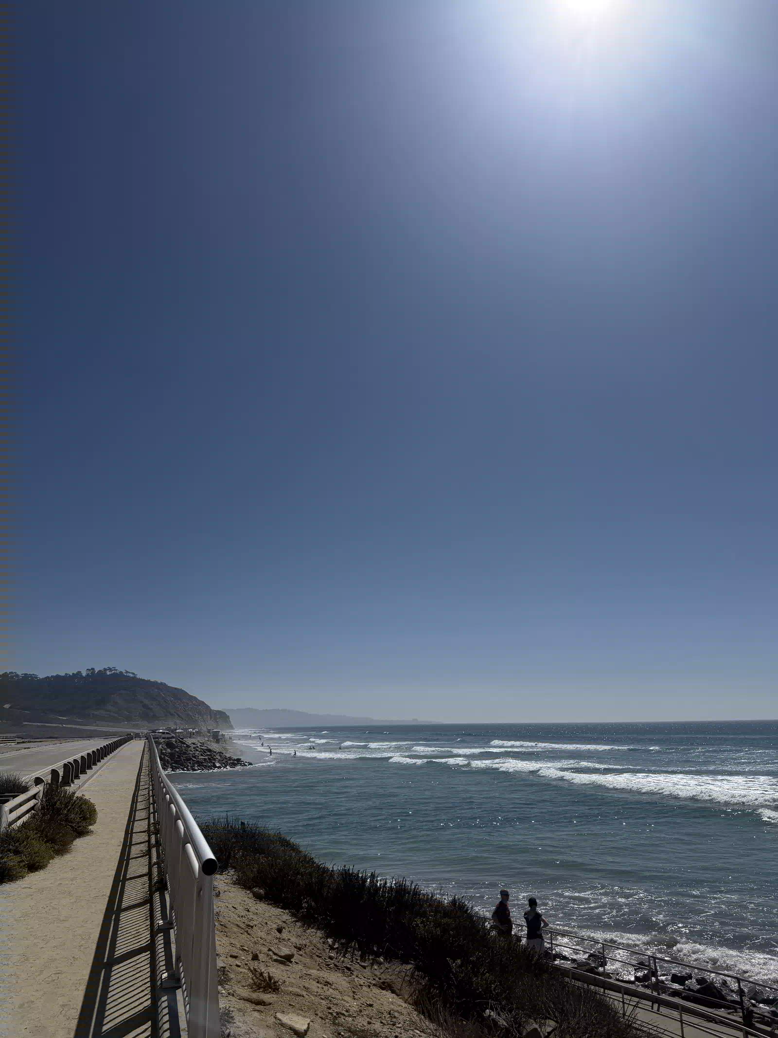 The coast path at Torrey Pines State Beach, a railing running ahead toward the bluffs, surf on the right and the sun high in a deep blue sky