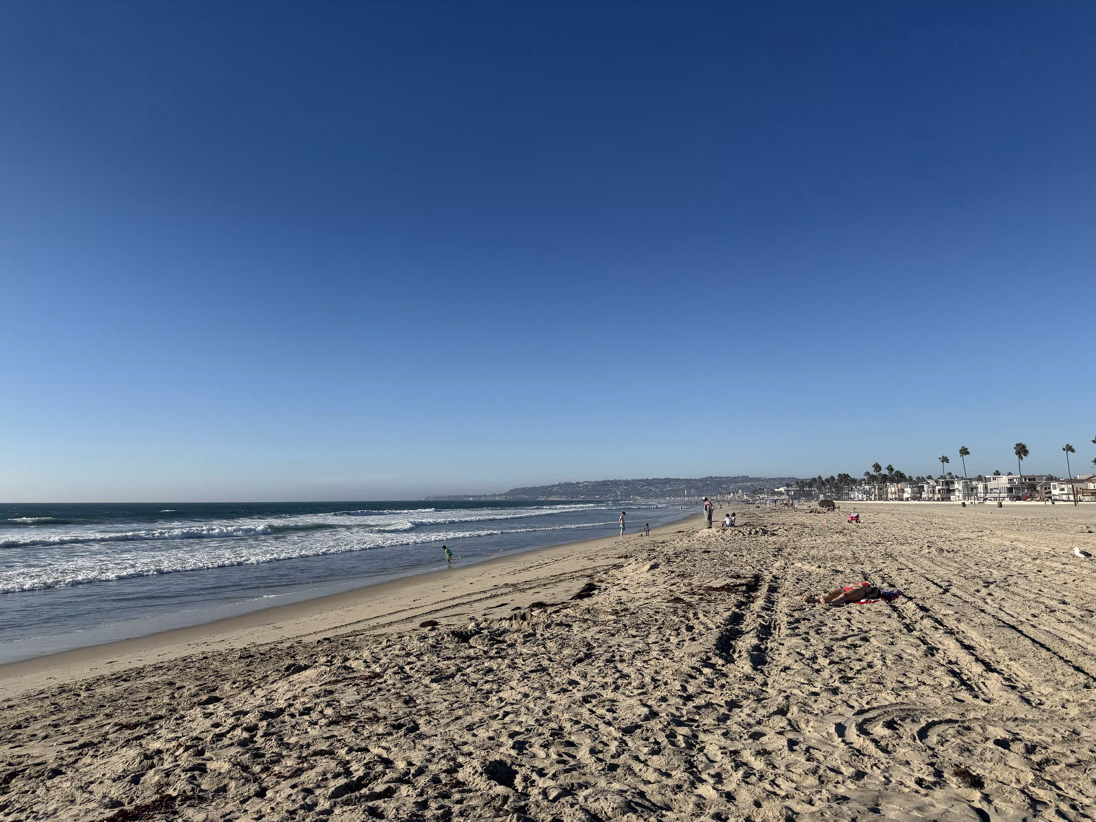 Mission Beach in late afternoon light, wide sand with tracks across it, a few people at the waterline and La Jolla on the hill in the distance